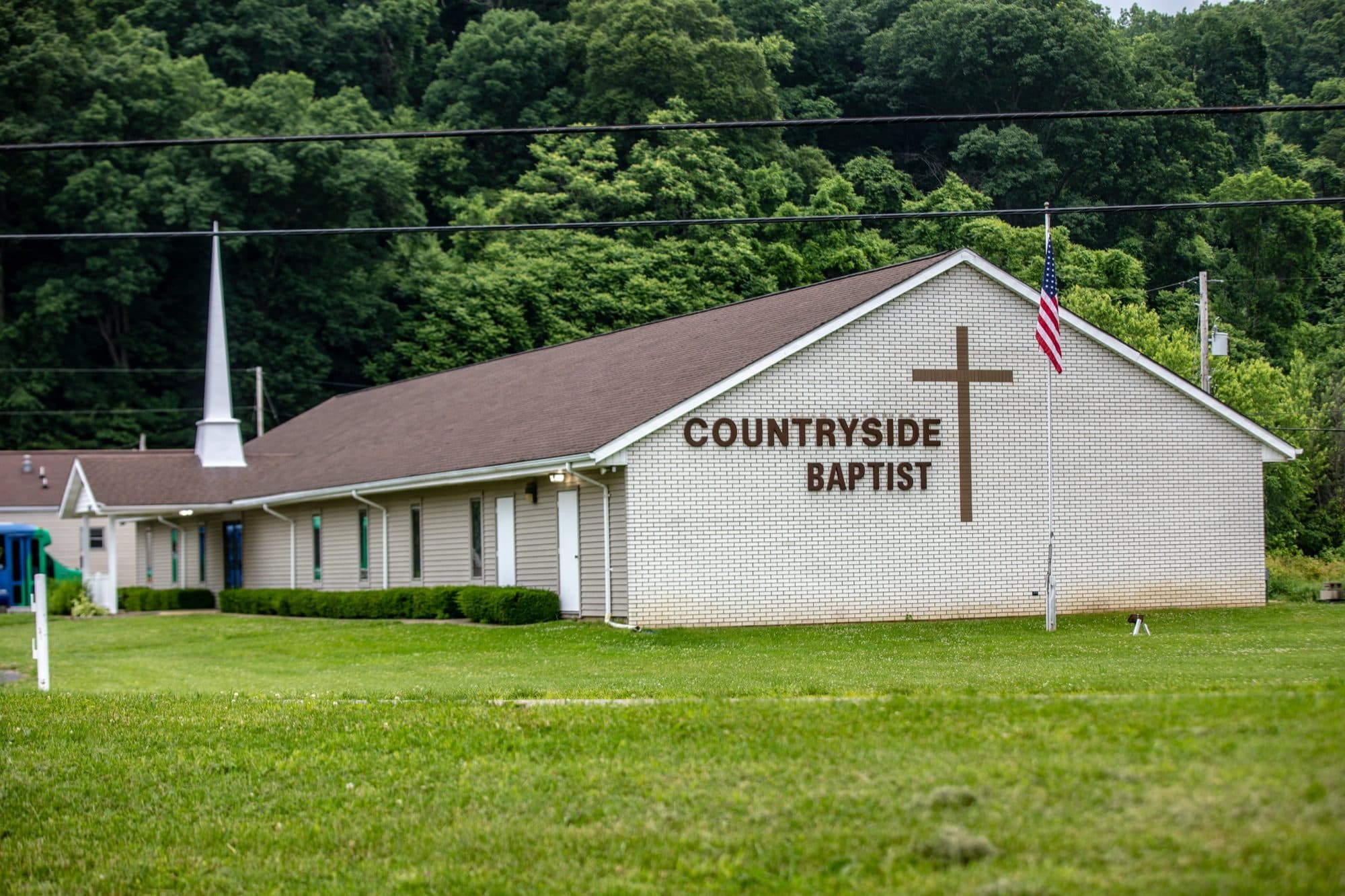Countryside Baptist Church seen from the road on a summer afternoon: a white brick gable carrying a tall cross and the words COUNTRYSIDE BAPTIST, a white steeple over the low entrance wing, an American flag on the pole, and a wooded hillside rising behind the building.