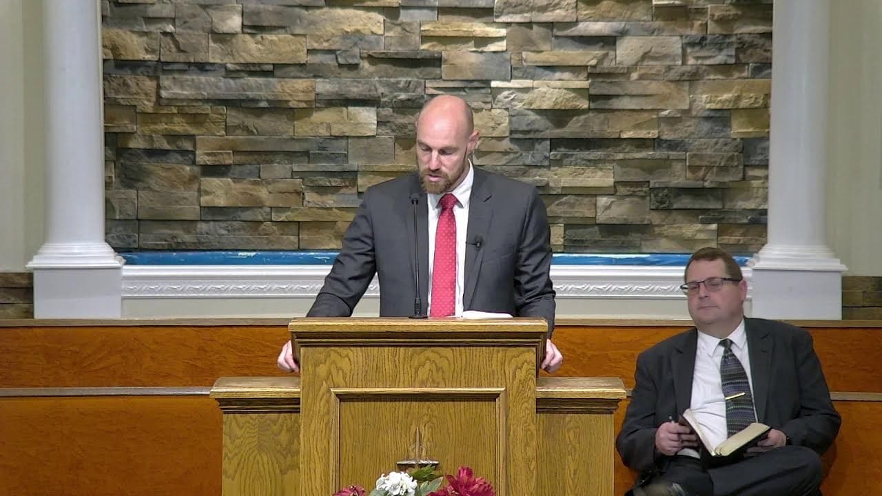 A guest speaker preaching at Countryside Baptist Church while Pastor Harvey listens from the platform bench with an open Bible.